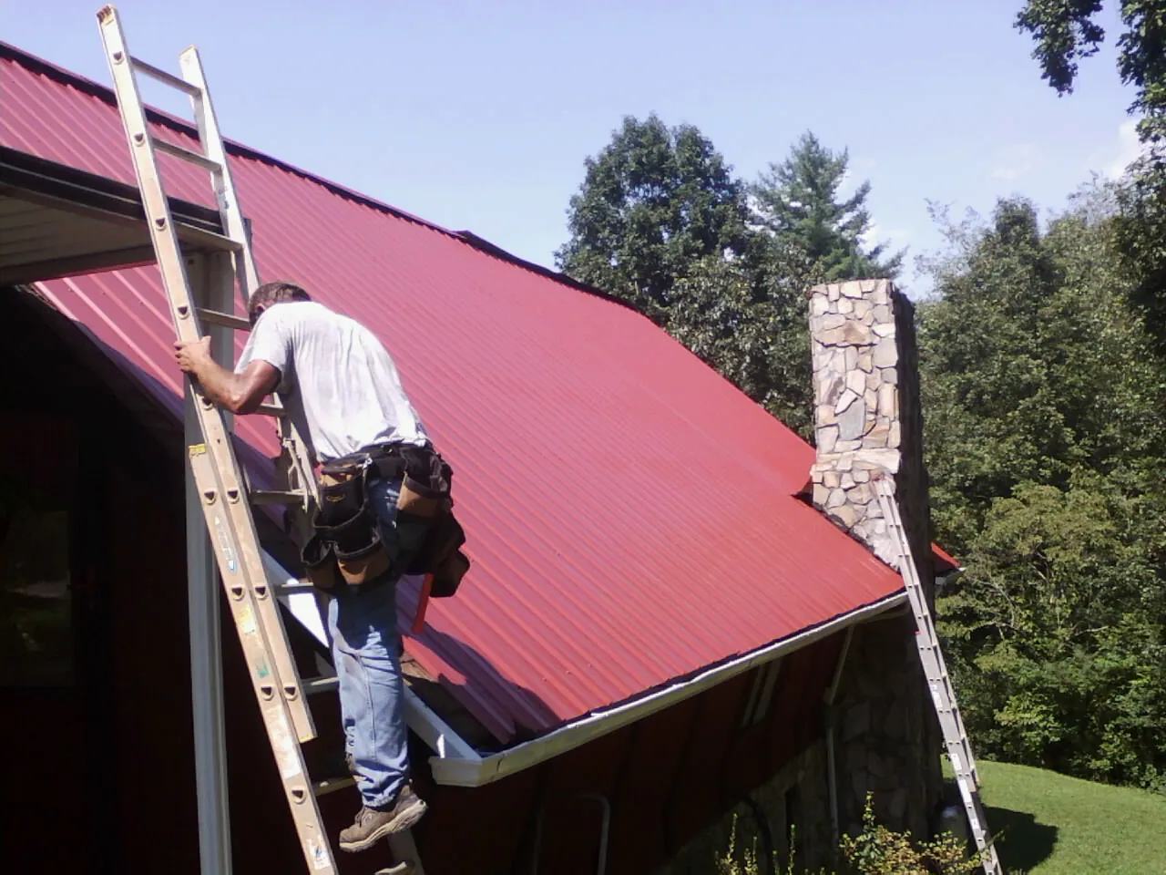 Roofer on a ladder beside a red metal roof and stone chimney — Soffit & Fascia Repair by Roofing Brothers in Liberal